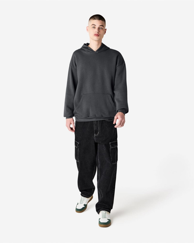 A unisex hooded sweatshirt in dark grey, worn by a model with baggy black trousers and white sneaker accents.