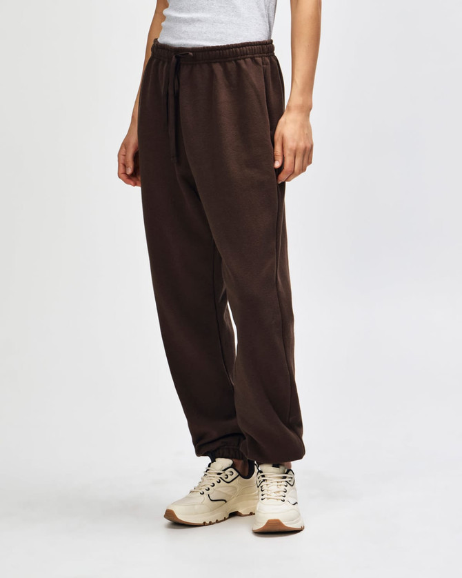 Brown unisex sweatpants with a drawstring waist, featured alongside black sneakers. The pants have a logo.