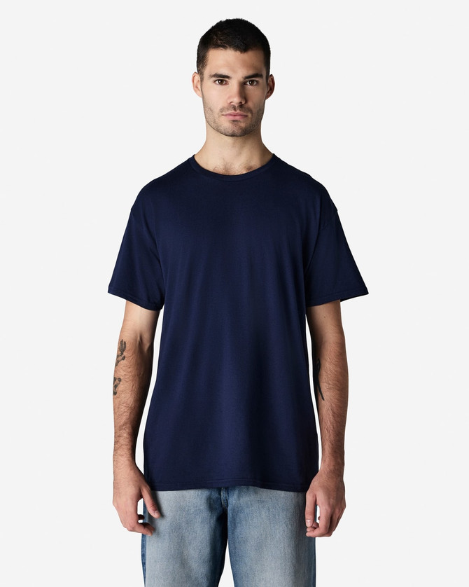 A man stands wearing a navy blue Gildan Light Cotton T-Shirt, paired with light blue jeans. The shirt has a logo.