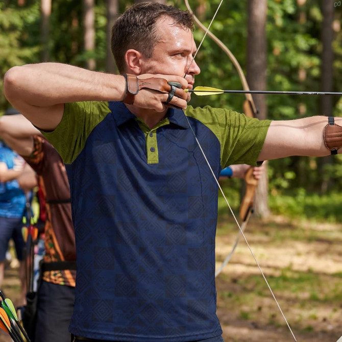 A man in a navy and green polo shirt is aiming an arrow with a bow in a forest setting. The shirt has a logo.