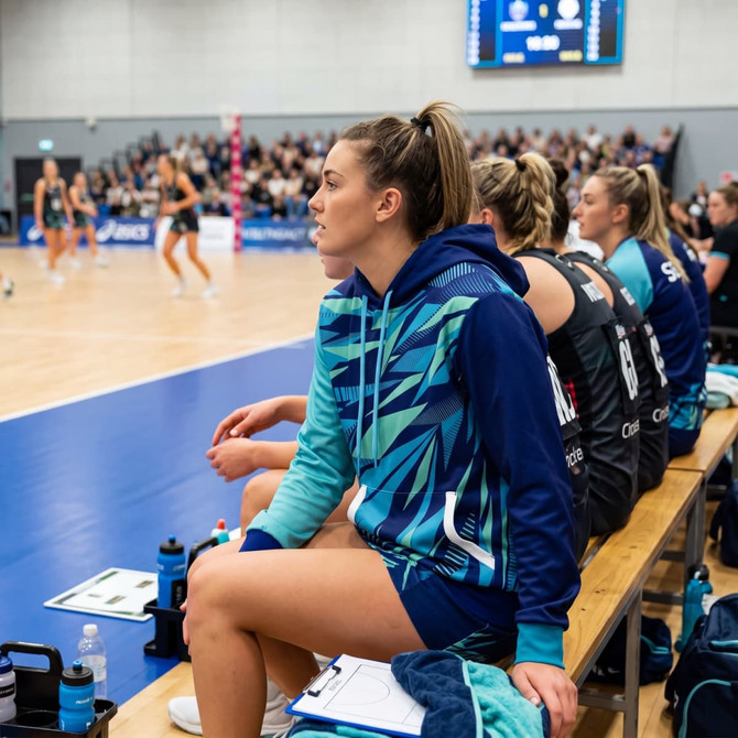 A custom women's active hoodie in teal and navy, featuring a logo, worn by a player on a sports bench.