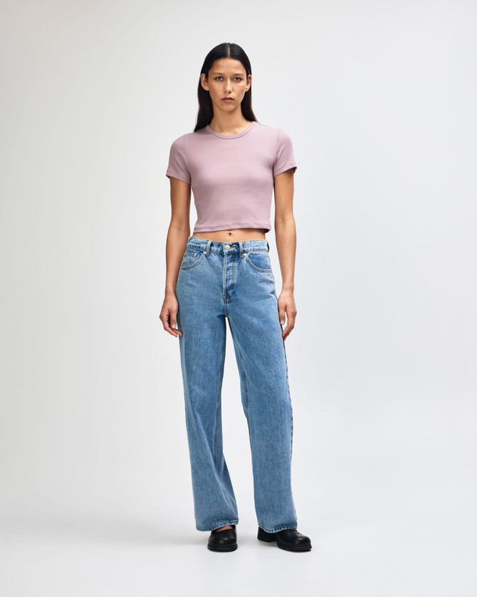 A fitted women's crop T-shirt in soft pink, paired with high-waisted blue jeans. The model stands against a light background.