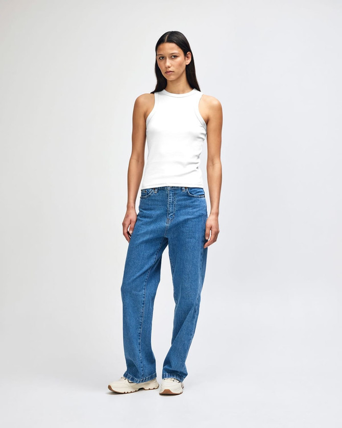 A white fitted tank top worn by a model, paired with blue jeans and casual shoes, standing against a light backdrop.