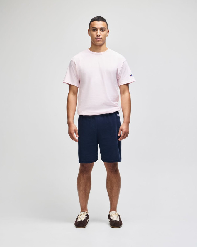 A man stands wearing a pink t-shirt and navy cotton gym shorts, featuring a logo on the shorts.