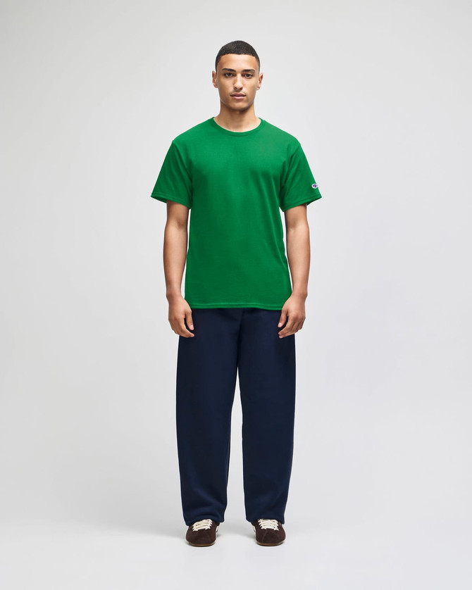 A green Champion adult jersey short sleeve t-shirt displayed on a model with navy blue trousers. The shirt features a logo.
