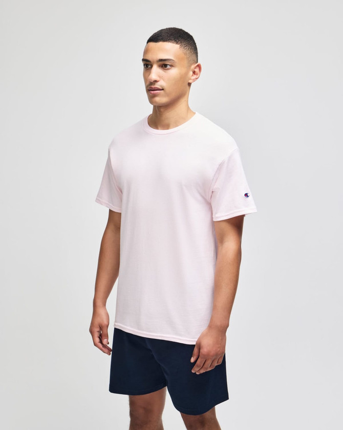 A pink Champion adult jersey short sleeve t-shirt displayed on a model, paired with navy shorts. The shirt features a branding logo.