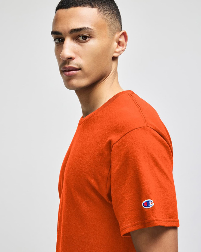 An orange Champion adult jersey short sleeve t-shirt displayed on a model. The shirt features a logo on the sleeve.