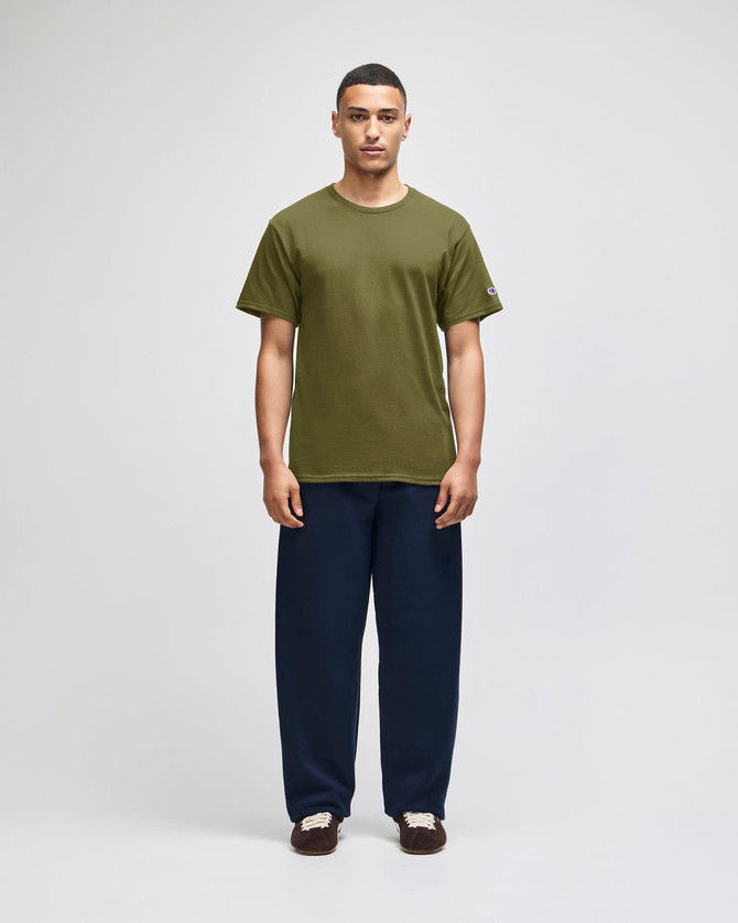 A model wearing an olive green short sleeve t-shirt and navy blue pants. The t-shirt features a logo.
