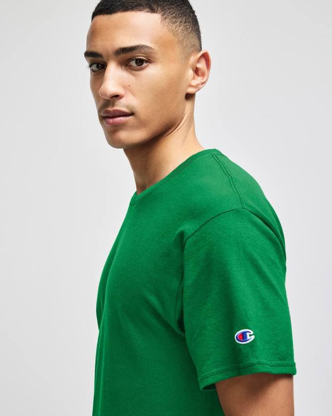 A green Champion adult jersey short sleeve T-shirt displayed on a model, featuring a logo on the sleeve.