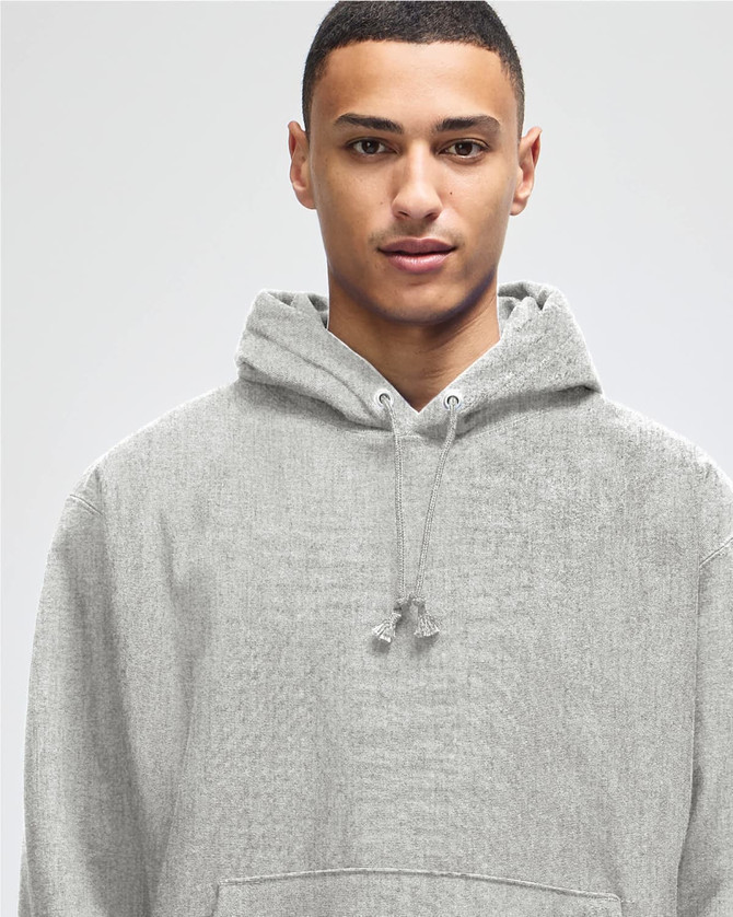 A grey hooded sweatshirt with a drawstring hood, featuring a logo. The model has short dark hair.