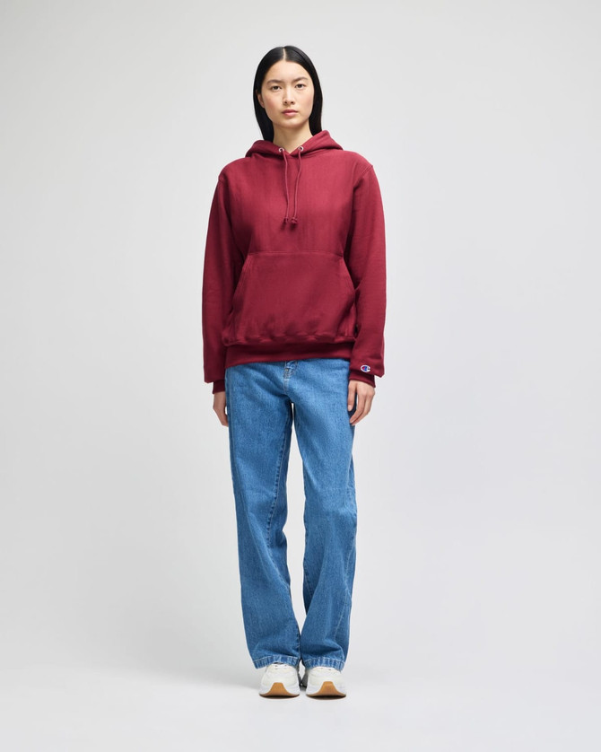 A model wearing a burgundy pullover hooded sweatshirt paired with light blue jeans, featuring a logo.