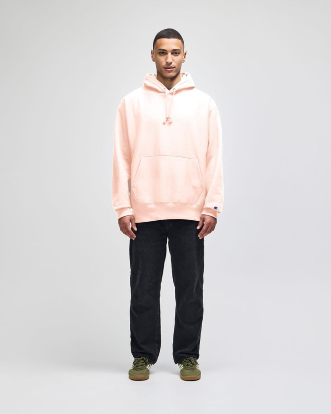 A person standing in a light pink hooded sweatshirt and black pants, wearing green shoes. The sweatshirt features a logo.