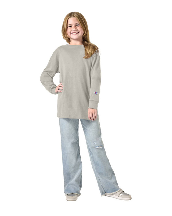 A youth long sleeve t-shirt in grey, paired with light blue jeans, featuring a logo on the sleeve.