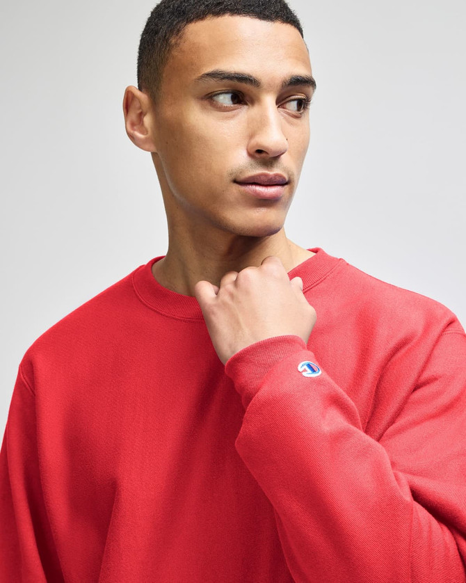 A red sweatshirt featuring a logo on the sleeve, modelled by a person looking to the side.