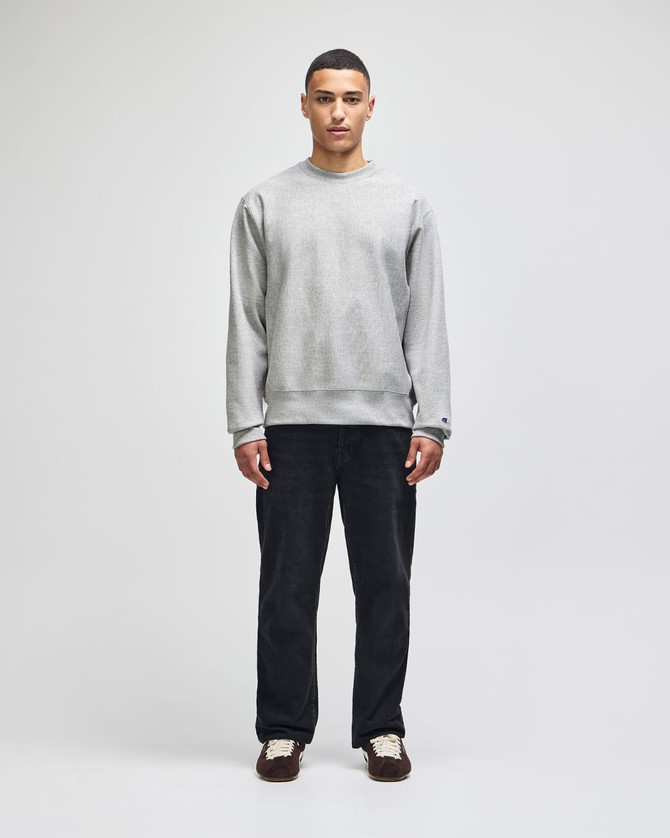 A grey crew sweatshirt worn by a model, paired with black trousers, featuring a subtle logo.