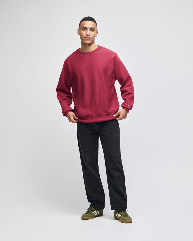A red crew sweatshirt is worn by a model standing in a casual pose, paired with dark trousers and green shoes.