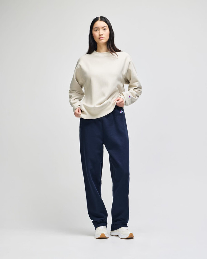 A model wears a beige Champion Reverse Weave® adult crew sweatshirt paired with navy trousers. The sweatshirt has a small logo.
