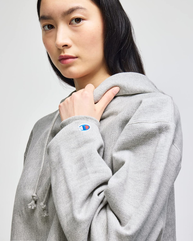 A grey cropped hoodie with a large hood and a logo on the sleeve, modelled by a woman.