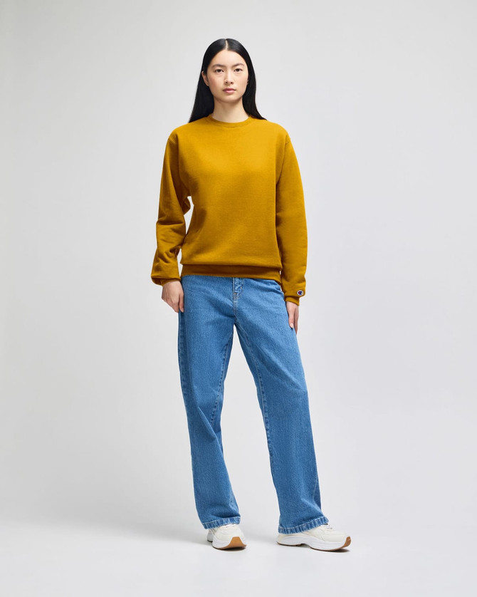 A model wearing a mustard yellow crewneck sweatshirt paired with light blue jeans and white shoes, featuring a logo.