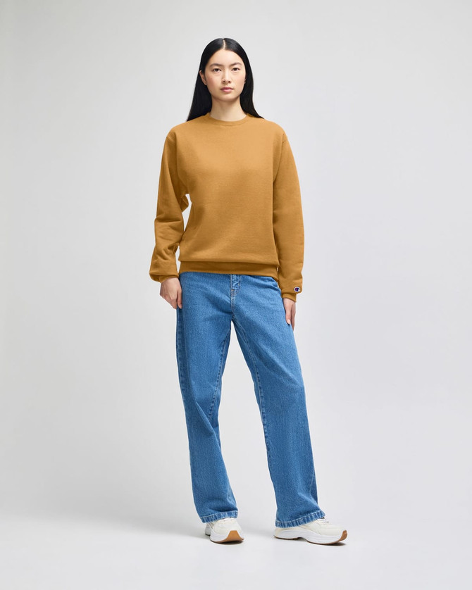 A model wearing a mustard-coloured sweatshirt and blue jeans, standing against a plain background. The sweatshirt features a logo.