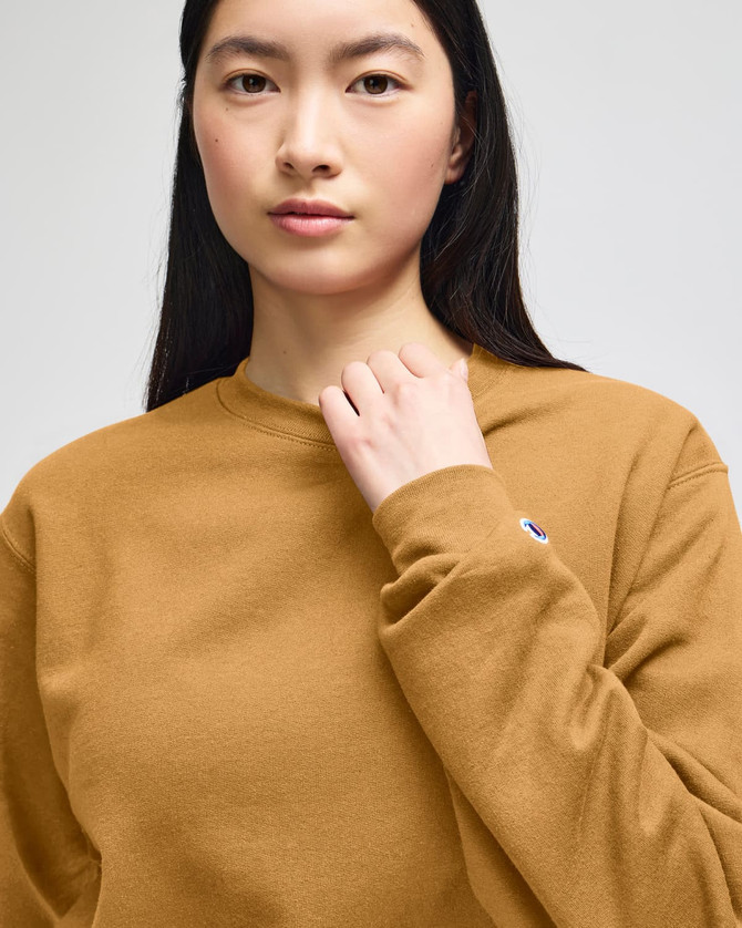 A mustard-coloured crewneck sweatshirt with a logo, worn by a model with long black hair.
