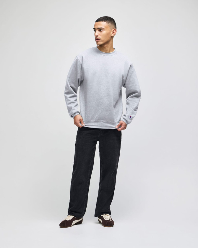A grey crewneck sweatshirt worn by a model, paired with black pants and shoes, featuring a logo on the garment.