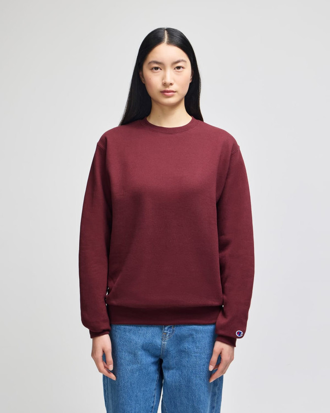 A maroon crewneck sweatshirt on a model, featuring a logo and styled with light blue denim jeans.