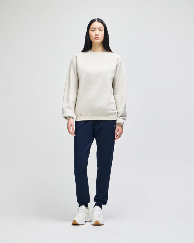A woman stands in a light grey Champion Powerblend® adult crewneck sweatshirt and navy jogger pants, wearing white sneakers.