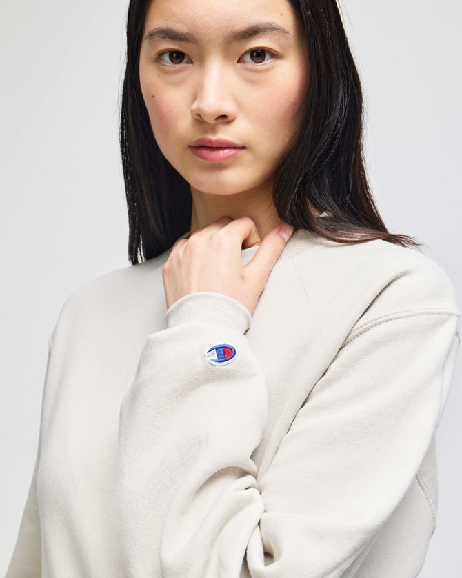 A lightweight, cream-coloured crewneck sweatshirt featuring a logo on the sleeve.