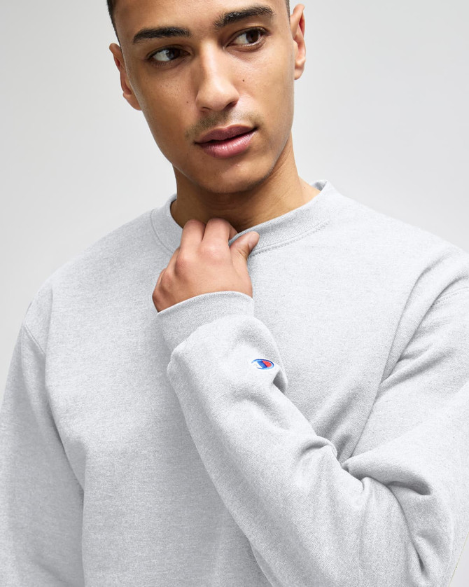 A grey crewneck sweatshirt on a model, featuring a logo on the sleeve. The fabric looks soft and comfortable.