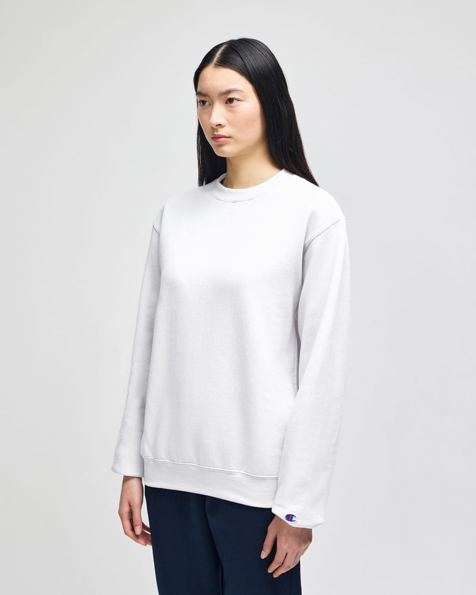 A white crewneck sweatshirt worn by a model, featuring a simple design and a logo on the sleeve.