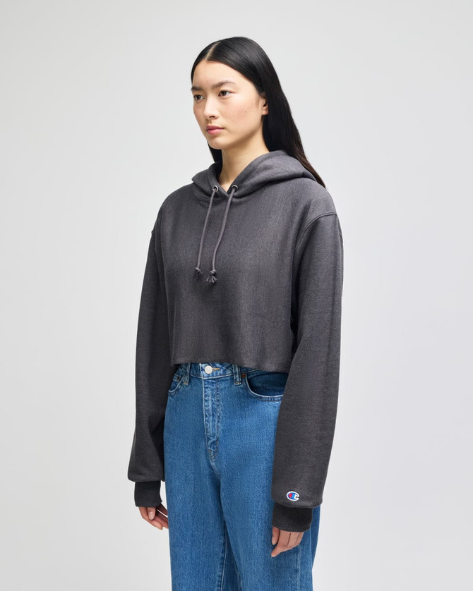 A model wearing a cropped grey hoodie with a hood and long sleeves, paired with blue jeans. The hoodie features a logo.