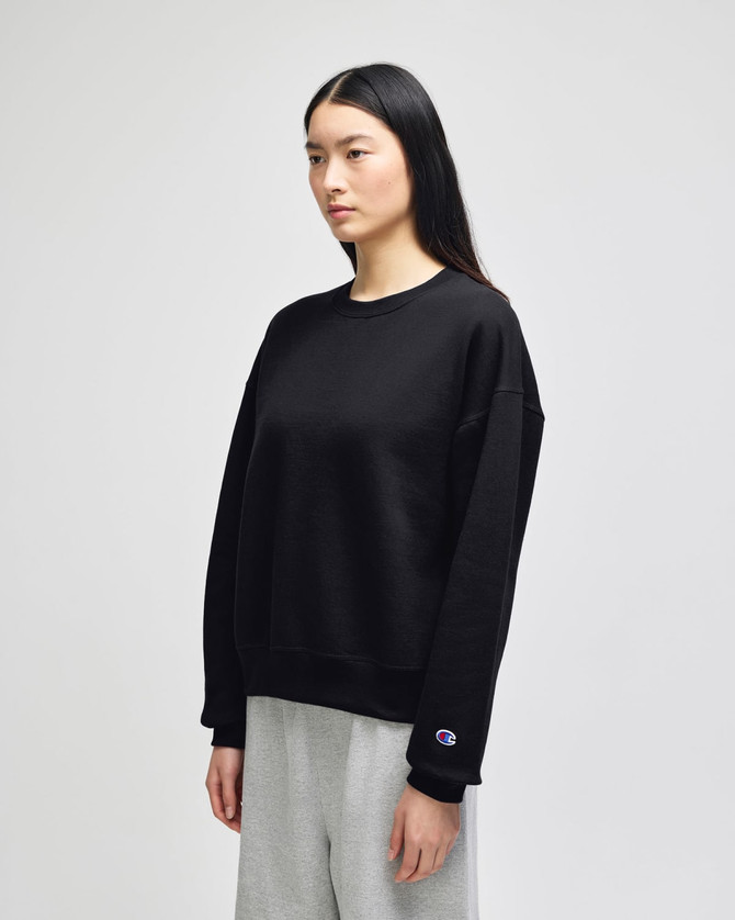 A black crewneck sweatshirt is displayed on a model, featuring a logo on the sleeve. The model wears grey pants.