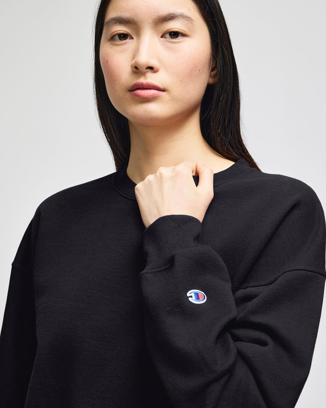 A black women's crewneck sweatshirt with a logo on the sleeve. The model is posing with a serious expression.