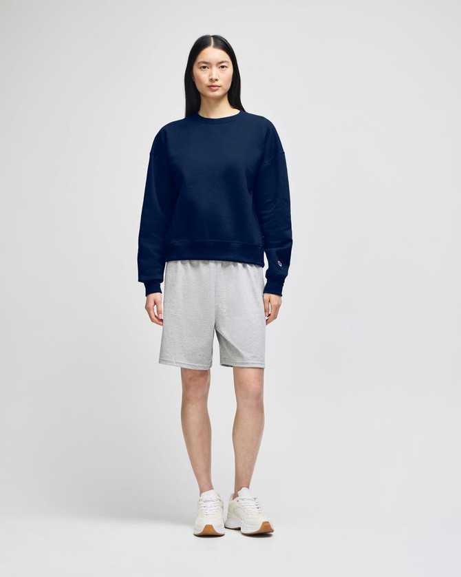 A navy crewneck sweatshirt worn with grey shorts and white sneakers, featuring a logo on the sweatshirt.