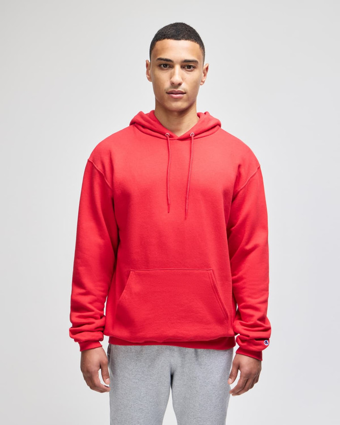 A red pullover hooded sweatshirt is worn by a model, styled with grey sweatpants. The garment has a front pocket and a logo.