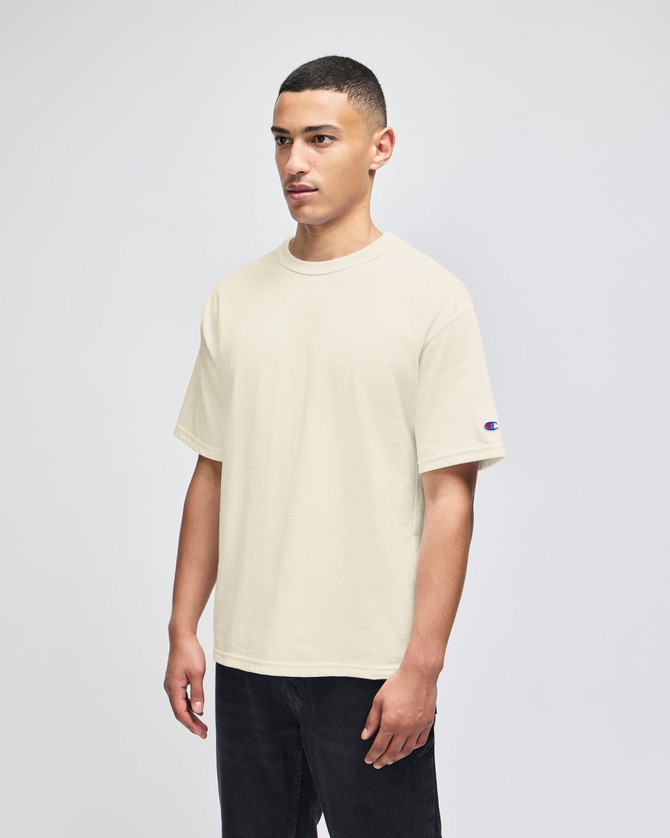 A beige Champion Adult Heritage Jersey T-Shirt on a model, featuring a logo on the sleeve.
