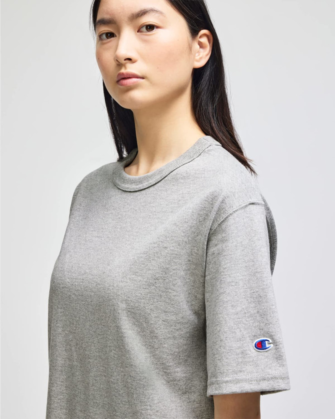 A grey Champion Adult Heritage Jersey T-Shirt is displayed on a model, featuring a logo on one sleeve.
