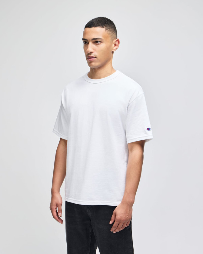 A white Champion Adult Heritage Jersey T-Shirt on a model, featuring a logo on one sleeve.