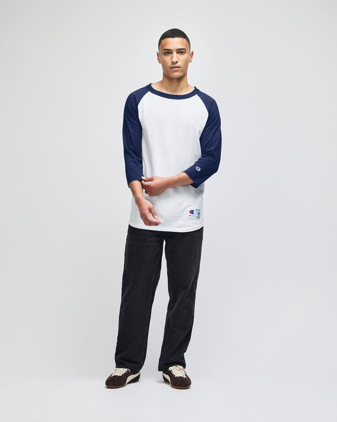 A model wearing a white and navy raglan baseball T-shirt, paired with dark pants and shoes. The shirt has a logo.
