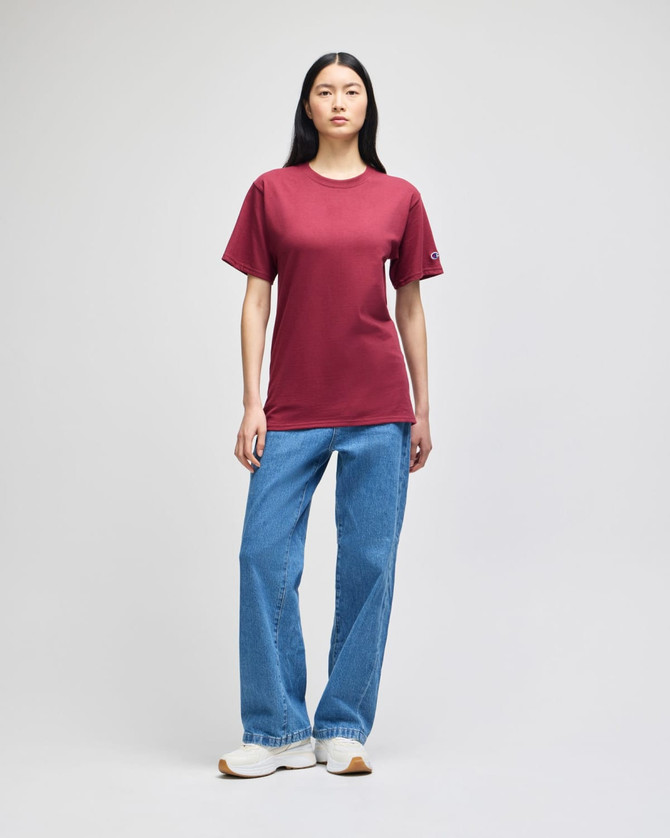 A woman is wearing a burgundy short-sleeve t-shirt paired with blue jeans and white sneakers. The shirt features a logo.