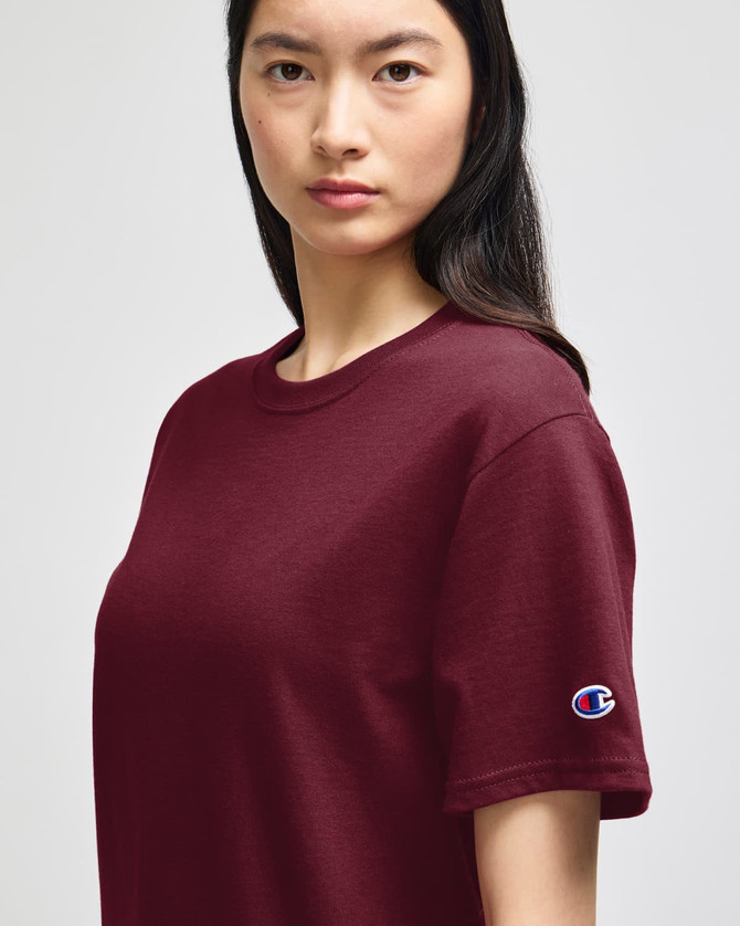 A maroon short-sleeve t-shirt featuring a logo on the sleeve. The model presents the shirt in a neutral setting.