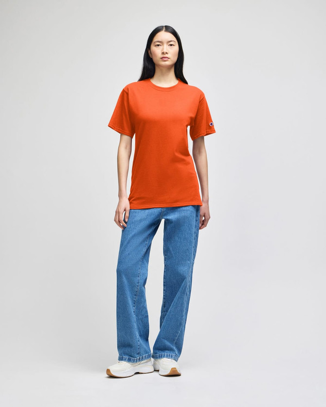 An orange short-sleeve t-shirt worn by a model, paired with blue jeans and white sneakers. The shirt features a logo.