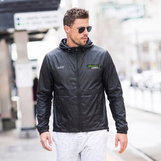 A man wearing a black windbreaker with a hood, paired with sunglasses and light-coloured trousers. The jacket has a logo.