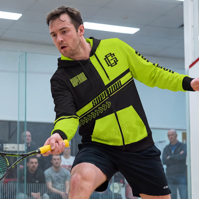A man wearing a black and bright green zipped hoodie is playing squash. The hoodie features a logo and sporty design.