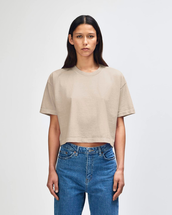 A cropped boxy t-shirt in a neutral beige colour, featured on a model wearing blue jeans. The shirt has a logo.