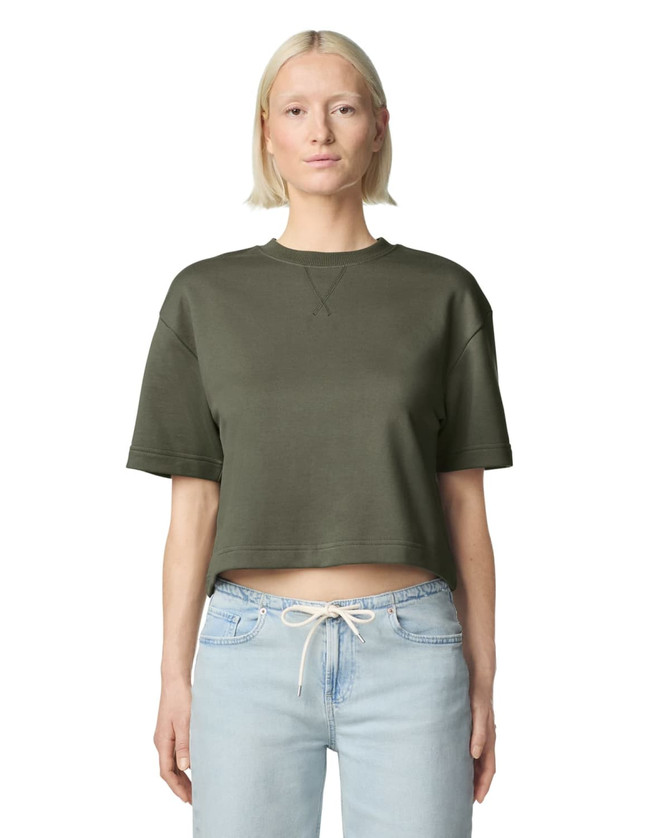 A woman wearing an olive green mid-length boxy top with short sleeves, paired with light blue jeans. The top features a logo.