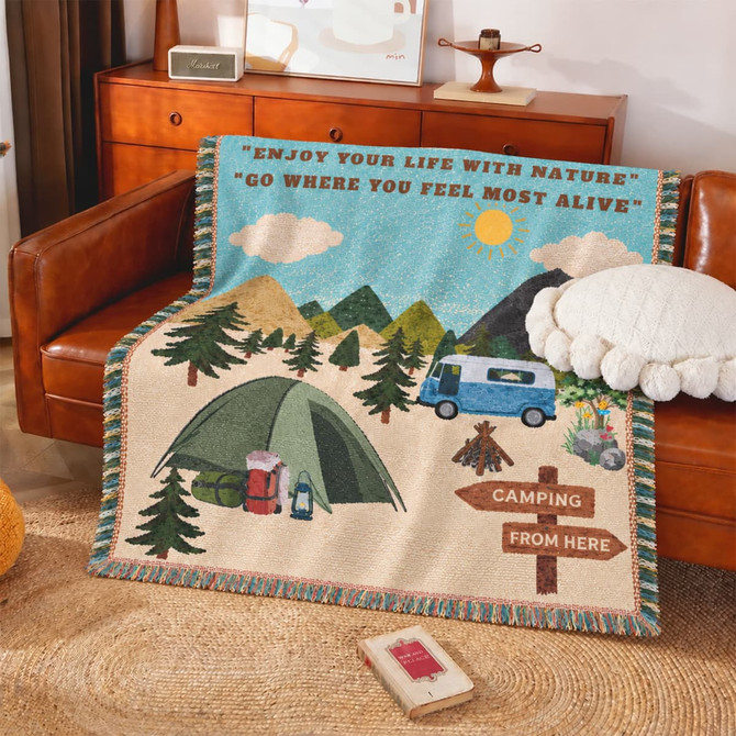 A tapestry blanket featuring a camping scene with mountains, trees, a tent, and a caravan in green and blue hues.