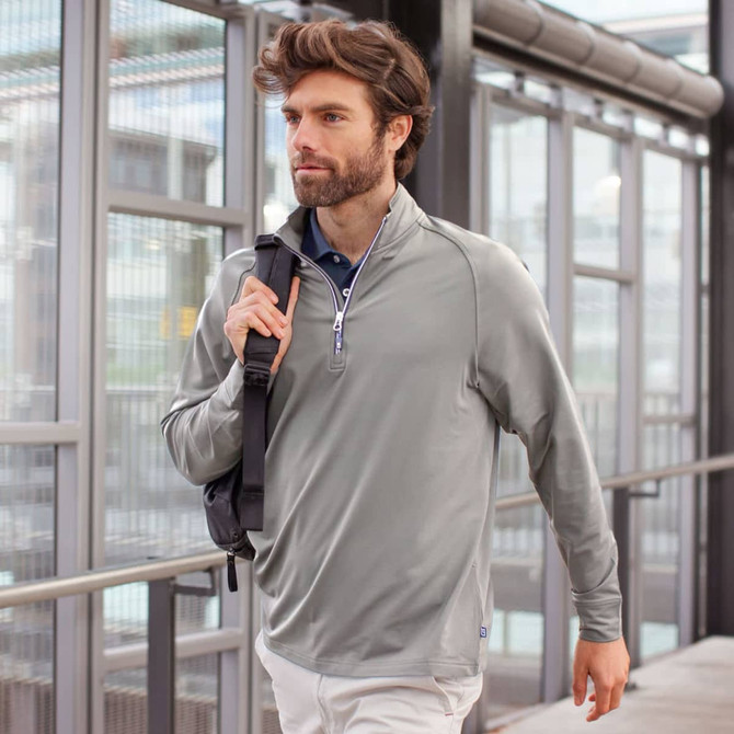 A man wearing a light grey half-zip sweatshirt, carrying a shoulder bag, walking in a modern building.