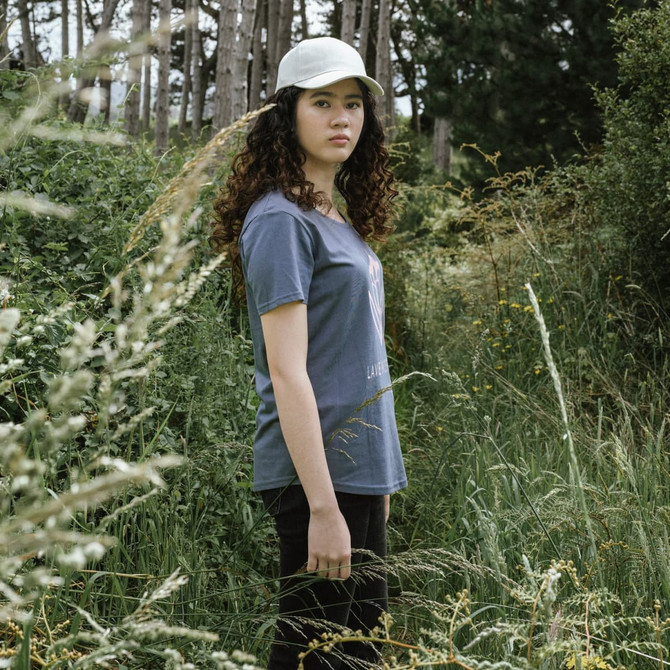 A young woman wearing a grey TRENDSWEAR Carmen women's T-shirt and a white cap stands in a grassy field.